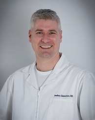 The image shows a man wearing a white lab coat, smiling and posing for the photograph.