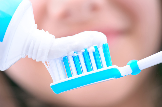 The image shows a close-up of a blue and white toothbrush with bristles being squeezed out, held by a human hand.