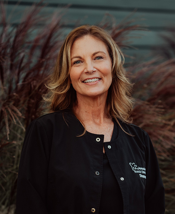 The image shows a woman standing outdoors, smiling at the camera. She has blonde hair and is wearing a black apron with white text on it. Behind her is a building with a sign that reads  Cafe  and there are plants visible in the background.