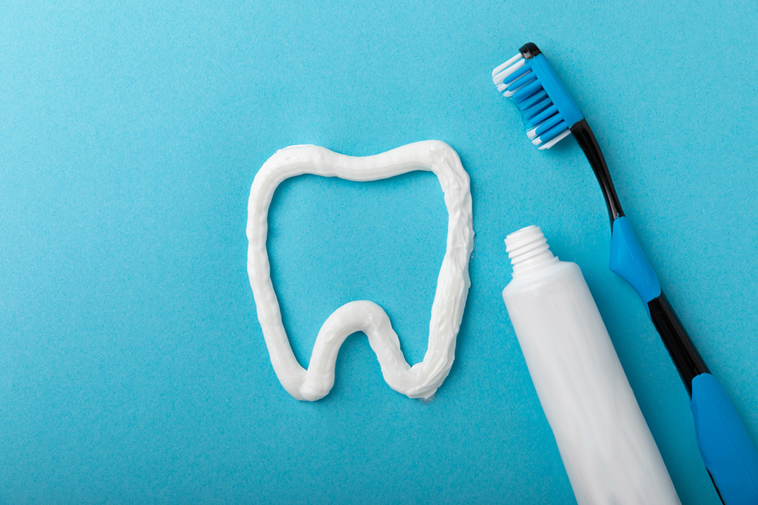 A blue toothbrush with a white handle and tube of toothpaste next to an illustration of a tooth on a light blue background.