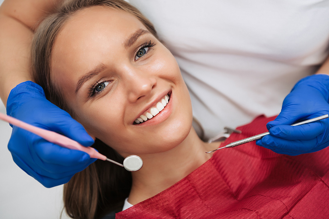 A woman is smiling while receiving dental care, with a dental hygienist working on her teeth and wearing protective gloves and a blue face mask.