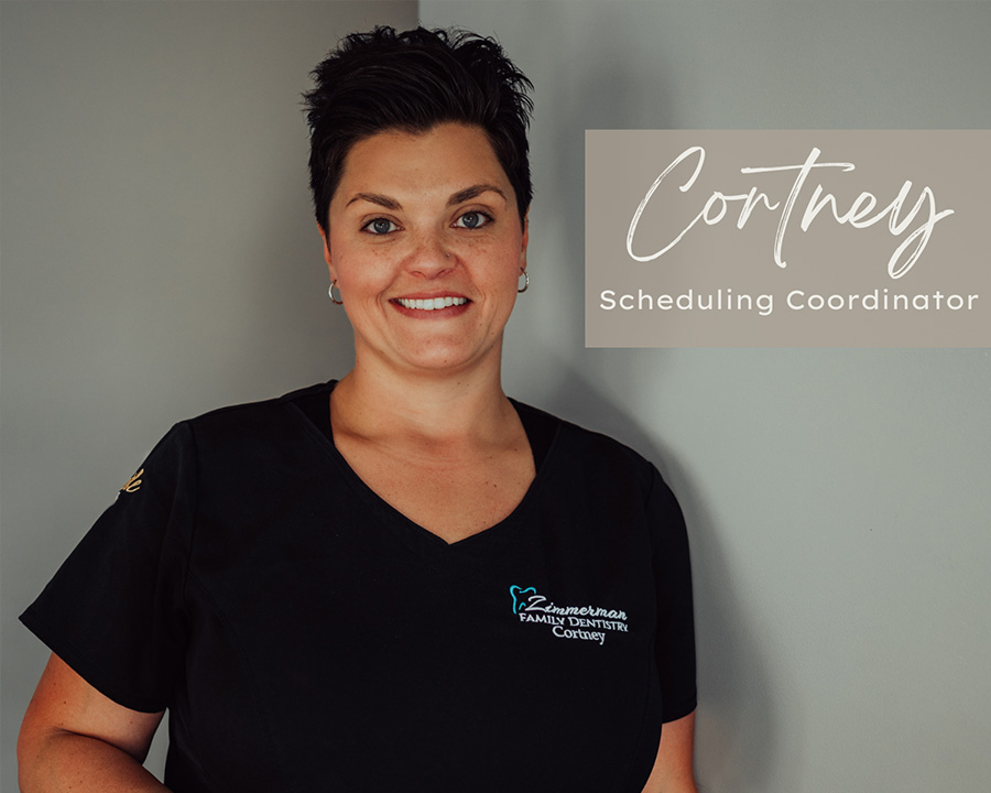 The image is a photograph of a woman standing against a light background. She has short hair, wears glasses, and is dressed in a dark top with a name tag that reads  Cortney.
