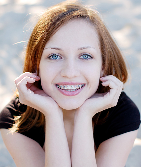 The image features a young woman with blue eyes and braces, posing for the camera with her hands on her cheeks. She is wearing a black top and has blonde hair.
