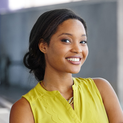The image depicts a smiling woman with dark hair, wearing a yellow top and standing in front of a building.