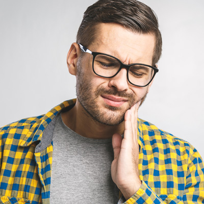 A man with a beard and glasses, wearing a yellow plaid shirt, is holding his hand to his mouth in a pained expression.