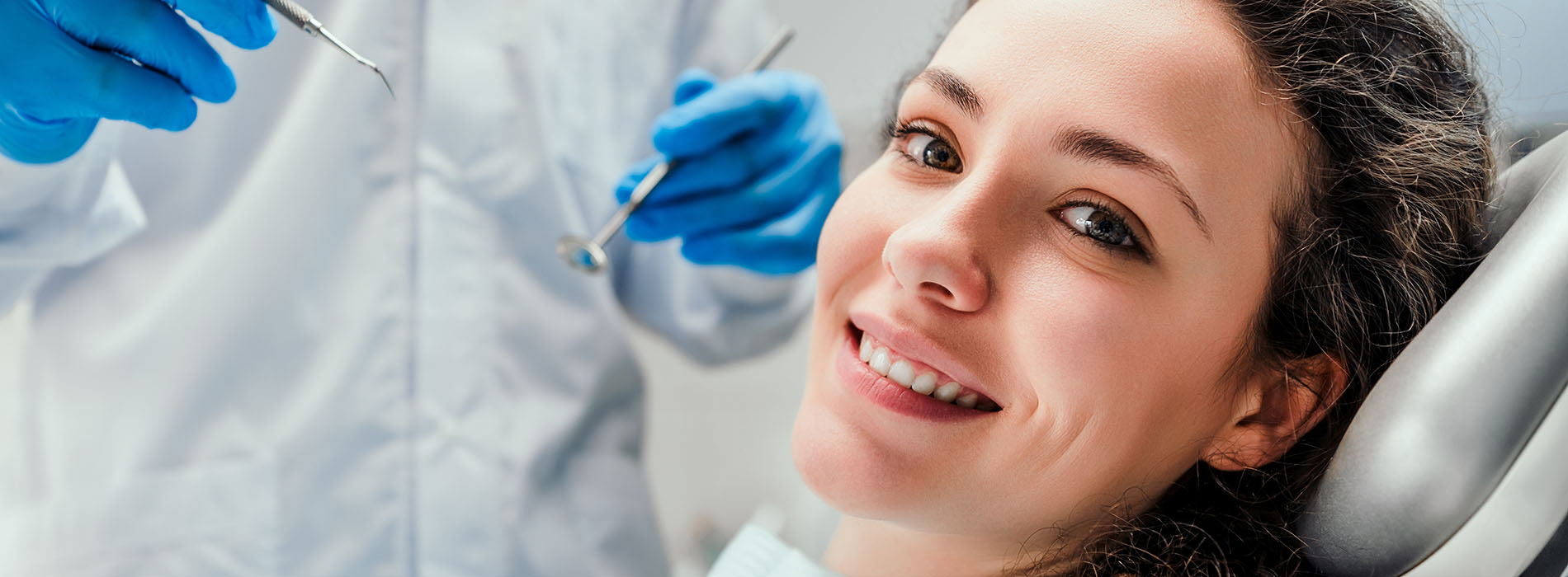 A woman is seated in a dental chair, receiving dental treatment with a smiling expression, while a dental professional works on her teeth.