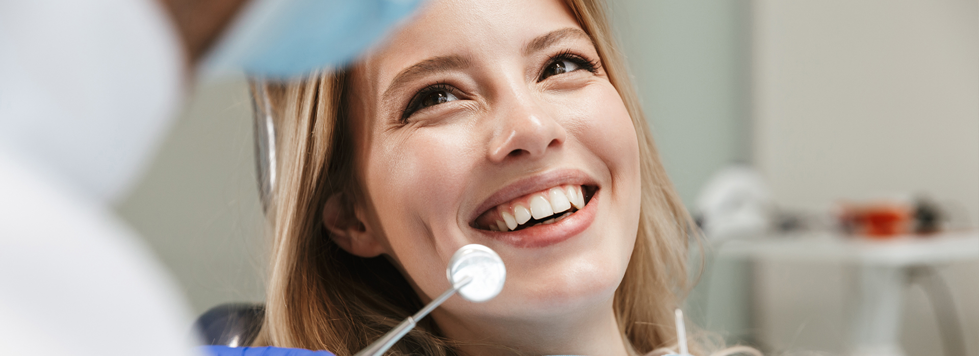 The image shows a woman sitting in front of a dental chair with a dentist, smiling and looking directly at the camera.