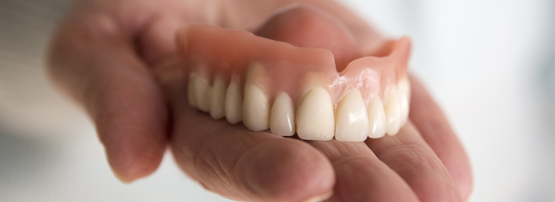 The image shows a human hand holding up a set of dentures, with the teeth visible against a blurred background.