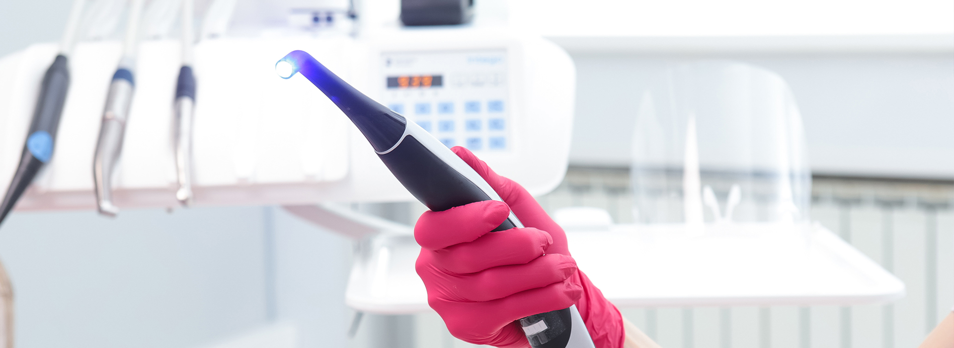 A person in pink gloves holds a blue device, possibly an ultrasonic cleaning tool, in front of a medical or dental setting.