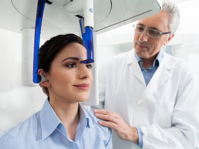 A woman in a blue shirt receiving a medical examination at a dental clinic, with a dentist standing behind her, both under a large digital x-ray machine.