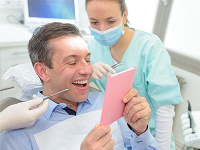 A dentist in a professional setting, holding a pink card and smiling at the camera while seated in front of a patient.