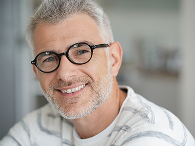 The image is a photograph of a smiling man with gray hair, wearing glasses and a white shirt, posing for the camera.