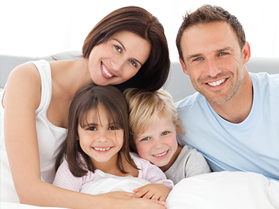 The image depicts a family of four, including an adult couple and two children, posing together on a bed with a smiling expression.