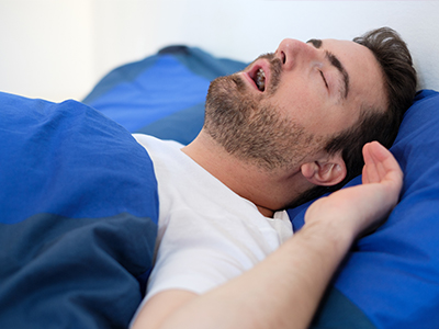 The image shows a man lying on his back in bed, with his eyes closed and mouth open as if he is sleeping or possibly snoring.