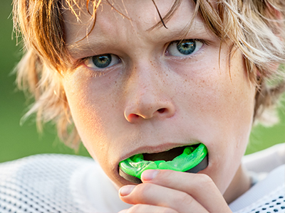 A young male athlete with blonde hair and a blue eye, holding a green sports mouthguard to his mouth.