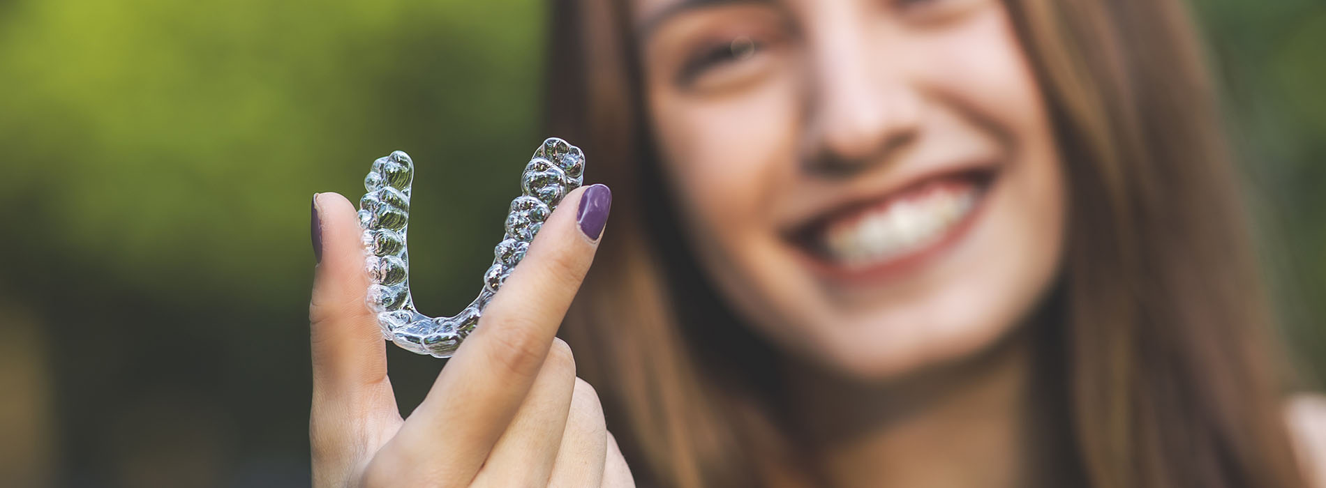 The image shows a person holding up a clear plastic dental retainer with their mouth partially open, smiling at the camera.